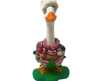 May include: A white goose figurine standing on a green base, wearing a red and white knit hat and a festive fabric outfit featuring horizontal stripes in red, green, and white, accented with small green pom-poms and a small green tree applique.
