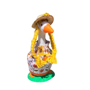 May include: A whimsical goose figurine wearing a straw hat and a floral dress. The goose has yellow yarn braids and an orange beak. It stands on a green base, creating a charming, decorative piece.