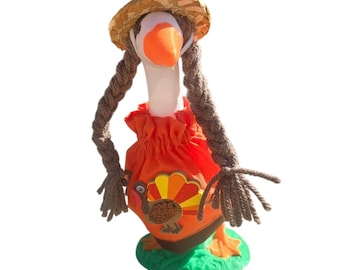 May include: A white goose figurine wearing a straw hat with two long brown yarn braids, dressed in an orange fabric outfit featuring a turkey applique on the front. The goose stands on a green base and has a bright orange beak and orange feet.