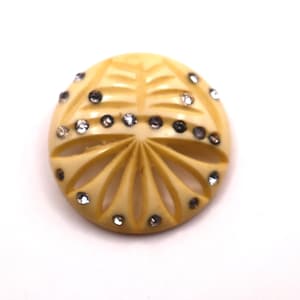Puede incluir: Un botón redondo de color crema con un diseño floral tallado. El botón está adornado con pequeños diamantes de imitación claros y gris oscuro alrededor del perímetro. El botón está sobre un fondo blanco.