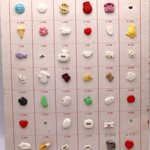 Puede incluir: Un tablero de exhibición vintage titulado "NYLON NOVELTIES 2" con una variedad de botones de plástico de colores. Los botones tienen varias formas, incluyendo un niño, un signo de dólar, un teléfono y un corazón. El tablero mide aproximadamente 30 cm por 25 cm.