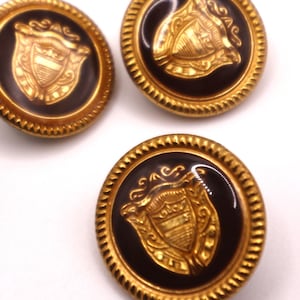 Puede incluir: Primer plano de varios botones redondos con borde dorado y centro marrón oscuro. Cada botón presenta un diseño de escudo dorado. Los botones tienen un borde texturizado en forma de cuerda.