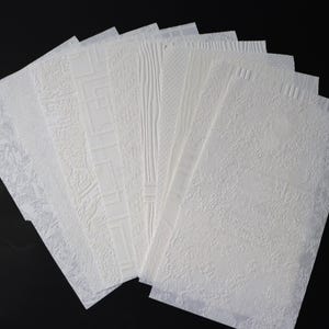May include: A collection of rectangular white sheets arranged in a fan shape, each featuring a distinct embossed texture. The patterns include geometric lines, wood grain, organic swirls, and fine cross-hatching, all rendered in a matte white finish.