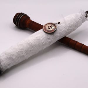 May include: Two wooden spools, one wrapped in white lace, are crossed against a white background. A decorative button secures the lace. The spools are dark brown with a textured design on the ends, suggesting a vintage or craft theme.