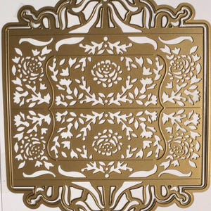 May include: A gold-colored metal craft die featuring an intricate rectangular frame with ornate bow motifs at the top and bottom. The central panel displays a detailed cut-out pattern of roses, leaves, and vines, creating a delicate openwork design.