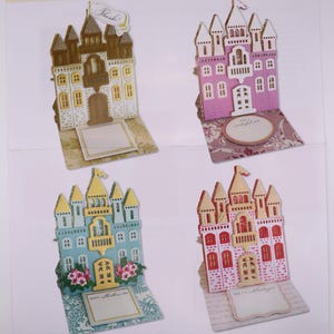 May include: An Anna Griffin Castle Easel Die instruction sheet featuring four distinct castle-shaped card designs in gold, purple, blue, and pink. Each castle includes intricate cutouts, turrets, and a base with a blank area for writing messages.