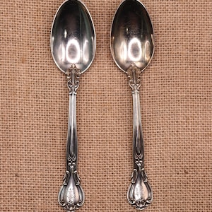 May include: Two antique silver spoons with decorative handles and oval bowls. The spoons have a reflective surface and are set against a neutral background. The handles feature detailed floral and scrollwork designs, adding a touch of elegance.