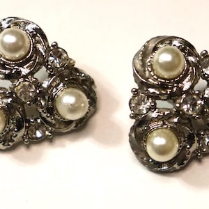 Puede incluir: Un par de pendientes plateados con cierre de clip. Cada pendiente presenta tres perlas de imitación y varios brillantes transparentes y brillantes. El diseño es ornamentado y de inspiración vintage, con un motivo floral.