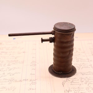 May include: A vintage brown metal oil can featuring a cylindrical, ribbed body with a flat circular base. It includes a long, thin horizontal spout extending from the top and a small plunger mechanism on the side for dispensing oil.