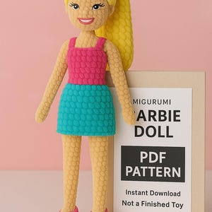 Può includere: Bambola amigurumi fatta a mano con capelli gialli a coda di cavallo, indossa un top rosa e una gonna turchese. La bambola ha scarpe rosa ed è accanto a un cartello con scritto "Amigurumi Barbie Doll PDF Pattern".