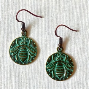 May include: A pair of antique-style earrings featuring a bronze-colored bee design. The earrings have a teal patina finish on the bee pendant and a dark brown hook. The round pendant is approximately 1 inch in diameter.