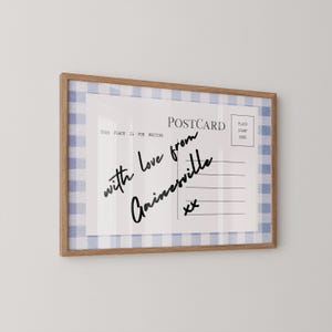 Pode incluir: Postal emoldurado com uma borda xadrez azul e branca. O postal diz "POSTCARD" e apresenta texto manuscrito que diz "with love from Gainesville" e "xx". A moldura &eacute; feita de madeira marrom clara.