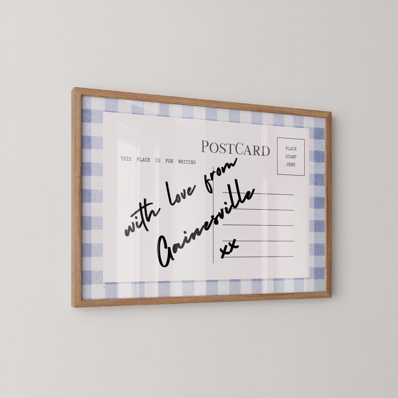 Pode incluir: Postal emoldurado com uma borda xadrez azul e branca. O postal diz "POSTCARD" e apresenta texto manuscrito que diz "with love from Gainesville" e "xx". A moldura &eacute; feita de madeira marrom clara.