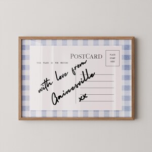 Pode incluir: Uma impress&atilde;o emoldurada de cart&atilde;o postal com uma borda de padr&atilde;o xadrez vichy azul claro e branco. O cart&atilde;o postal diz "with love from Gainesville" em escrita preta. A moldura &eacute; de madeira marrom clara.