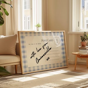 Pode incluir: Uma impress&atilde;o de postal emoldurada com uma moldura de madeira clara. O postal apresenta uma borda com padr&atilde;o xadrez azul e branco e o texto manuscrito "with love from Gainesville xx". O postal est&aacute; encostado a um sof&aacute; bege.
