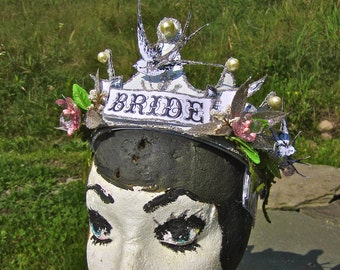 May include: A white and silver bridal crown with the word "BRIDE" on the front. The crown is decorated with white and silver feathers, white beads, and pink flowers.