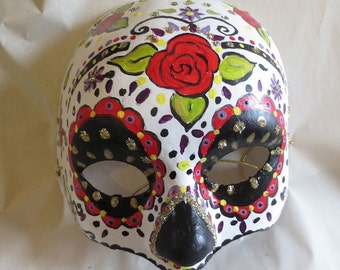 May include: A white skull mask with black eye sockets and a black nose. The mask is decorated with red, yellow, green, purple, and gold paint. The mask has a floral design with a large red rose in the center.