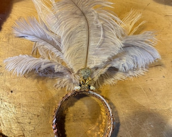 May include: A vintage-style headband with a brown sequined band and a cluster of light brown feathers. The headband features a decorative gold-colored centerpiece with small white embellishments, creating a glamorous accessory.