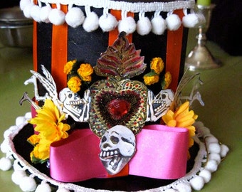 May include: A black and orange striped top hat with white pom-poms around the brim. The hat is decorated with a pink ribbon, a heart with a red jewel, two skeletons, and flowers.