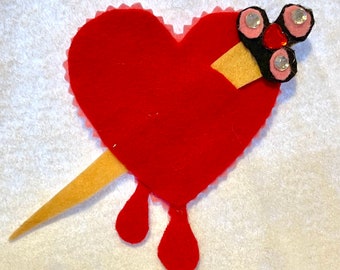 May include: A red felt heart with a pink and black felt arrow piercing through it. The arrow has three rhinestones on the tip. The heart has three red felt drops of blood dripping from the bottom.