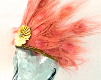 May include: A pink feather headdress with a gold and white decorative accent. The headdress is worn on a glass mannequin head.