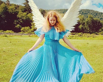 May include: A woman wearing a blue polka dot dress with white angel wings behind her. She is standing in a field with green grass and a blue sky in the background.