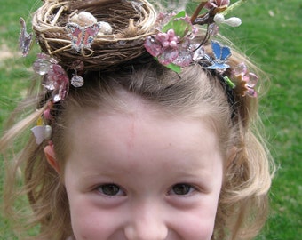 May include: A young girl wearing a whimsical hair accessory with a bird's nest, a bird, butterflies, and flowers. The accessory is made of natural materials and colorful embellishments.