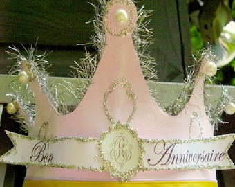 May include: A pink paper crown with silver tinsel and white beads. The crown has a white banner with the words "Bon Anniversaire" in black script.