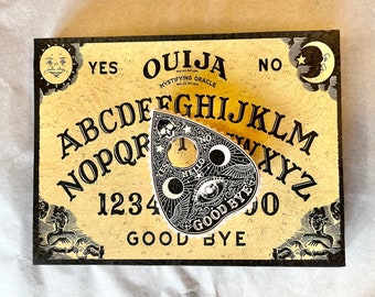 May include: A wooden Ouija board with a black planchette featuring the words "Hello" and "Good Bye". The board has a yellow background with black letters spelling out the alphabet, numbers 1-9 and 0, and the words "Yes", "No", and "Good Bye".