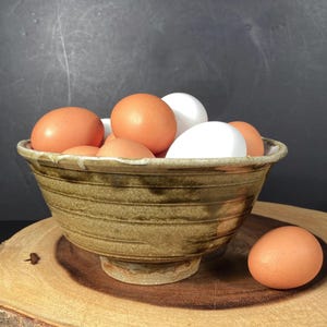 May include: A ceramic bowl filled with brown and white eggs, set on a wooden surface. The bowl has a textured, earthy appearance with horizontal lines. One egg rests outside the bowl. The background is a dark, neutral color.