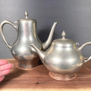 May include: Two antique silver-colored teapots. One is tall with a curved handle and spout, the other is rounder with a shorter spout and handle. Both have lids with a pointed finial. The teapots are displayed on a wooden surface.