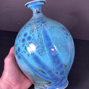 May include: A handcrafted ceramic vase with a turquoise glaze and dark blue accents. The vase features a bulbous body, a narrow neck, and decorative blue patterns. The vase is held in a hand.