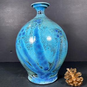 May include: A turquoise ceramic vase with a round body and a narrow neck. The vase features a textured surface with darker blue accents and a decorative pattern. A gold-colored pinecone sits beside the vase.