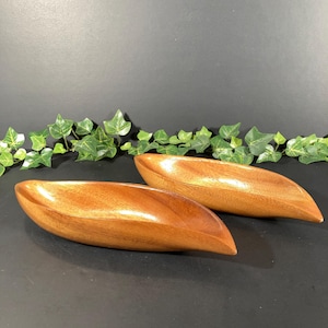 May include: Two wooden, boat-shaped bowls with a glossy finish. The bowls are a warm brown color and appear to be made of a hardwood. The bowls are empty and sit on a dark surface, with green ivy in the background.