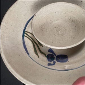 May include: A handmade ceramic bowl and plate set. The off-white pottery features a speckled texture and is decorated with blue and green floral designs. The bowl sits atop the plate, showcasing a rustic, artisanal aesthetic.
