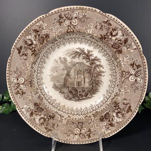 May include: A vintage brown and white decorative plate with a detailed floral and landscape design. The plate features a central scene of a building and figures, surrounded by intricate floral patterns and a scalloped edge. The plate is displayed on a clear stand.