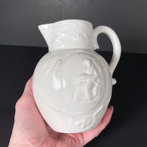 May include: A white ceramic pitcher with a rounded body and a curved handle. The pitcher features embossed wheat and a figure in a dress. The spout is pointed, and the pitcher is held in a hand.