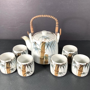 May include: A white ceramic tea set with a bamboo design. The set includes a teapot with a woven handle and six matching cups. The bamboo stalks are brown and the leaves are blue. The teapot has a white lid.