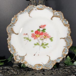 May include: A white, scalloped-edge plate with a floral design. The plate features a central cluster of pink flowers with green leaves. The rim is decorated with gold and light blue accents. The plate is displayed on a decorative metal stand.