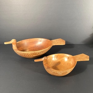 May include: Two wooden duck-shaped bowls. The bowls are carved from light brown wood, with a smooth finish. Each bowl has a duck head and tail, and the bowls have decorative carvings around the rim. The bowls are on a dark surface.
