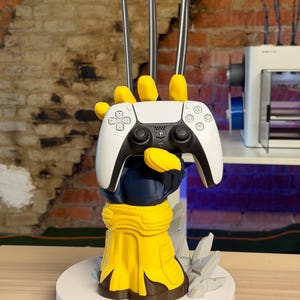 Pode incluir: Um suporte decorativo em forma de mão enluvada amarela e azul com três longas garras prateadas estendidas para cima. A mão segura um controle de PlayStation 5 branco e preto. A base é circular e apresenta detalhes cinzentos de rocha.