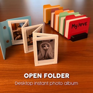 Pode incluir: Um álbum de fotos instantâneas de mesa com uma capa azul, exibindo várias fotos pequenas de um gato. Uma fileira de porta-fotos coloridos em forma de pasta também é visível. O texto "OPEN FOLDER" e "Desktop instant photo album" são exibidos.