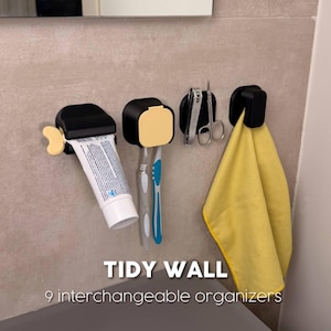 Pode incluir: Quatro organizadores de parede pretos com detalhes amarelos, com o texto TIDY WALL 9 interchangeable organizers. As unidades seguram um tubo de pasta de dentes, duas escovas de dentes, uma tesoura de metal e uma toalha amarela.