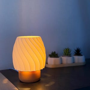 Pode incluir: Uma pequena luminária de mesa moderna com uma cúpula creme com padrão espiral e uma base canelada laranja-marrom. A lâmpada está acesa, emitindo um brilho quente. Três pequenas suculentas em vasos estão no fundo.