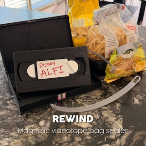 Pode incluir: Um selador de sacos magnético de fita de vídeo preta com a palavra "REWIND" e o texto "Magnetic videotape bag sealer" em branco. Uma fita VHS preta com a etiqueta "Disney ALFI" está em uma caixa preta aberta.
