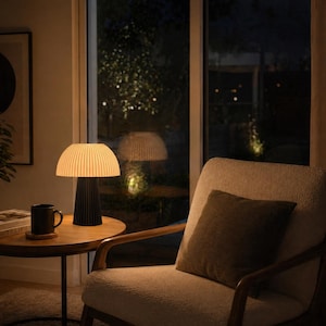 May include: A modern table lamp with a pleated lampshade and a black base sits on a round wooden side table. A black mug and a book are also on the table. An upholstered armchair with a brown pillow is next to the table.