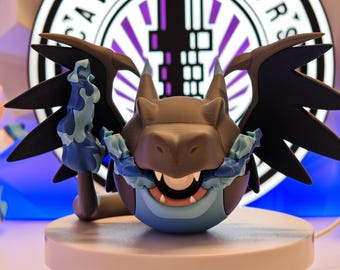 May include: A detailed figurine of a dragon-like creature with black wings, a grey head, and a blue belly. It has a wide grin and is holding a blue and white flame. The figurine is displayed on a white pedestal, with a sign that says "CAVE CREATORS" in the background.