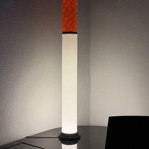 Könnte beinhalten: Eine hohe, zylindrische Stehleuchte mit einem weißen, lichtdurchlässigen Körper, der ein sanftes Licht abgibt. Der obere Teil ist in leuchtendem Orange gehalten und weist ein strukturiertes, mosaikartiges Muster auf. Sie steht auf einem schwarzen Fuß.