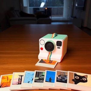 Pode incluir: Uma câmera instantânea branca e verde menta com um detalhe de listras arco-íris e um conjunto de oito porta-copos Polaroid. Os porta-copos apresentam várias imagens, incluindo paisagens e um gato. O texto "8 POLAROID COASTERS" é exibido.
