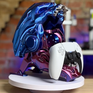 Pode incluir: Um suporte metálico azul e roxo para comando de videojogos em forma de cabeça de criatura com dentes afiados. O suporte tem um acabamento brilhante e iridescente e segura um comando sem fios branco e preto na zona da mandíbula.